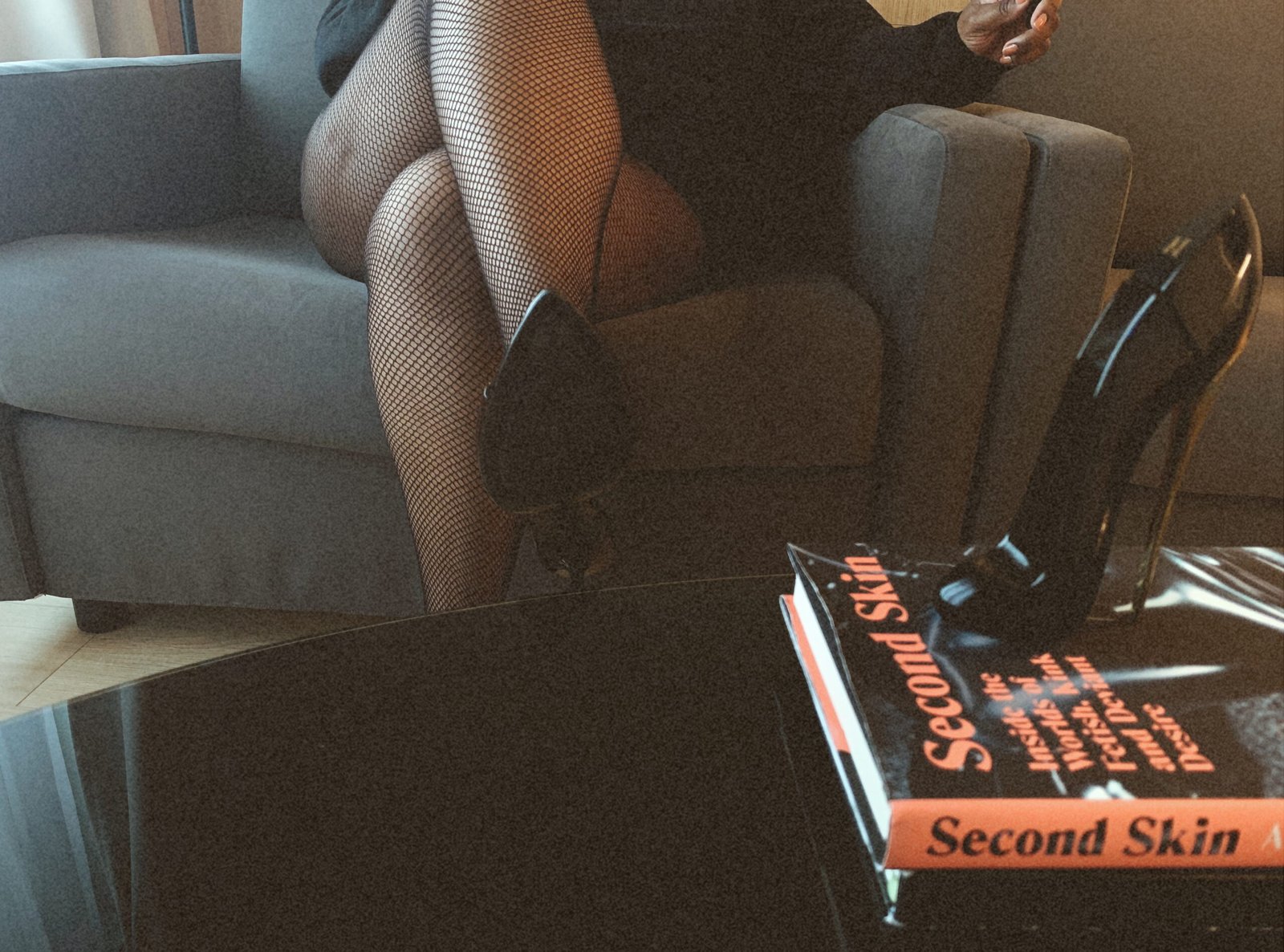 A pair of crossed legs in heels fishnets and heels sitting on a grey chair. Sable Gold, Luxembourg escort